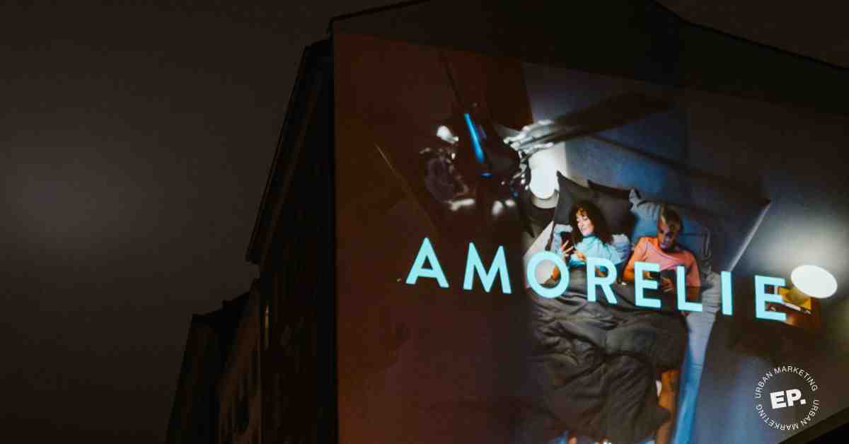 Großflächige Amorelie-Projektion auf einer Hausfassade bei Nacht: ein Paar liegt im Bett, beide am Smartphone. Eigene Umsetzung von Echo Poster.