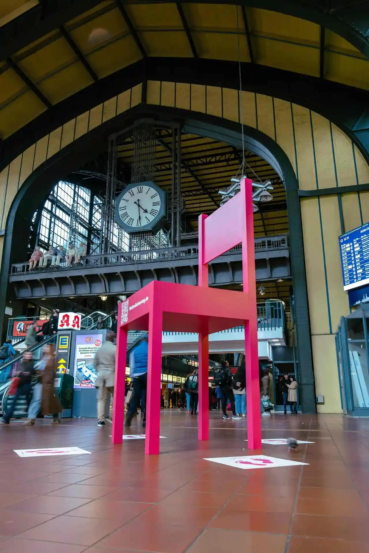 Überdimensionaler pinker Stuhl mit QR-Code im Hamburger Hauptbahnhof, auf dem Boden pinke Floor Graphics mit Fußabdrücken.